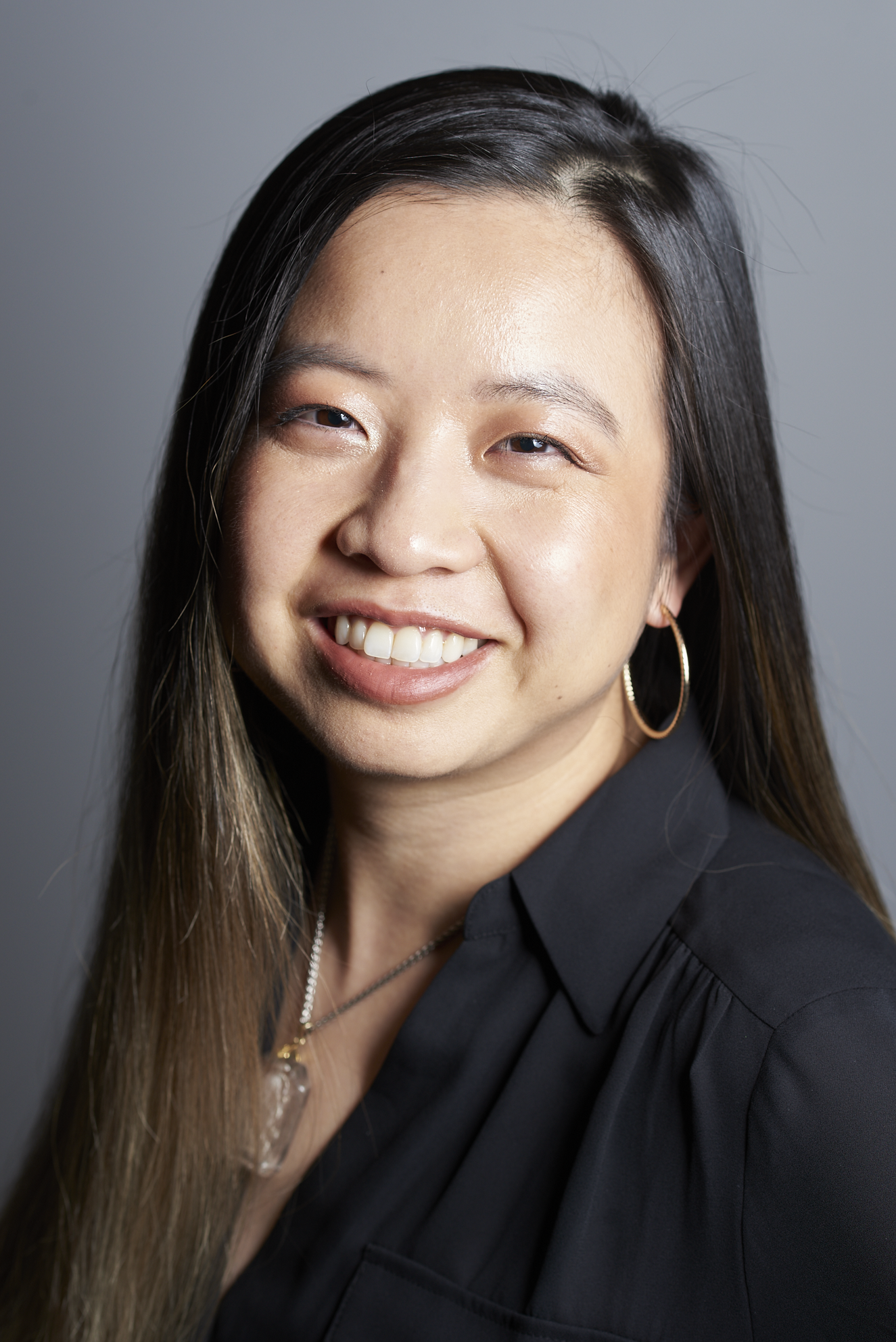 Close-up of a smiling woman with long, dark hair, wearing hoop earrings, a necklace, and a black top, against a gray background.