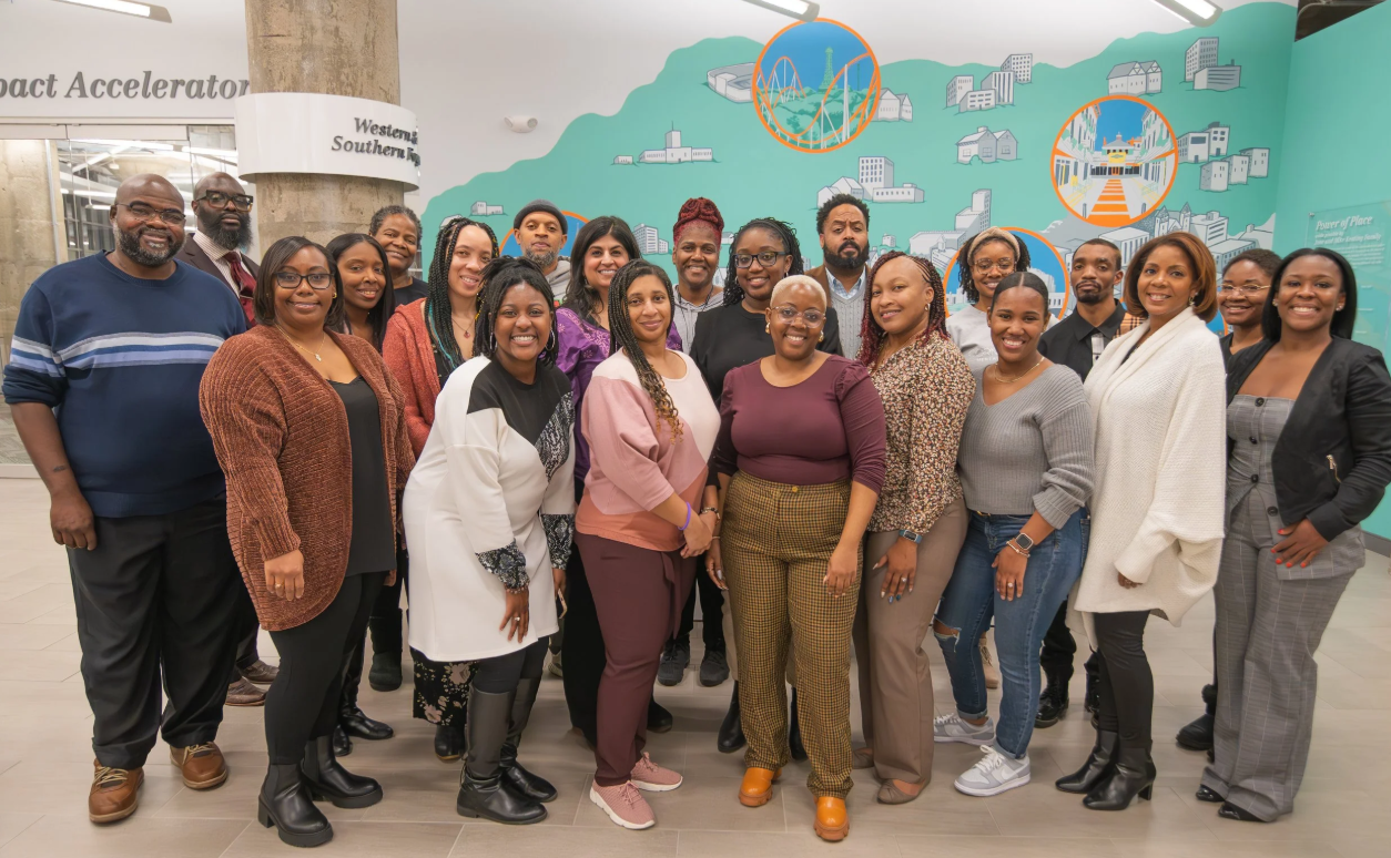 Group of diverse people posing for a photo in an indoor space with a mural backdrop of a cityscape and amusement park rides.