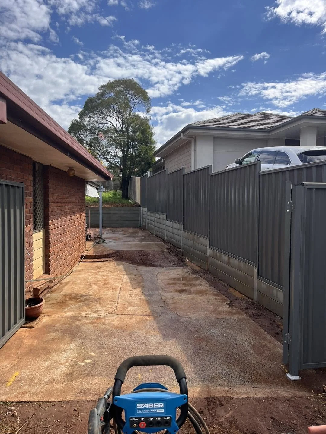 Backyard with concrete patio, a tall gray fence, and a small sloped yard with a tree in the background under a partly cloudy sky.