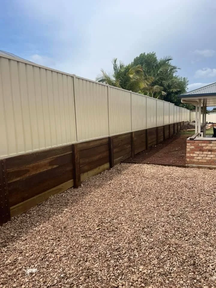 A backyard with a new privacy fence, some trees in the background, and a gravel ground surface near a brick structure with a covered area.