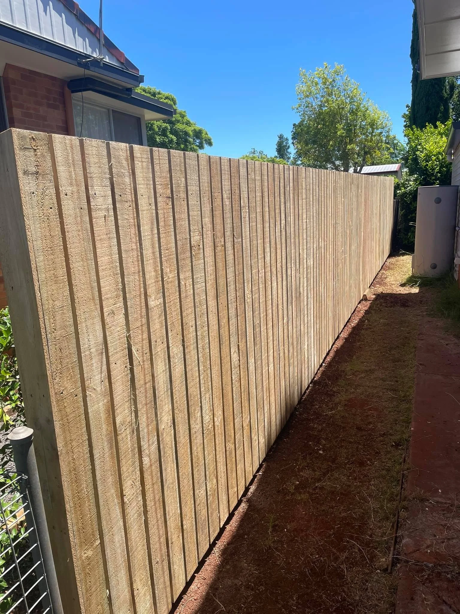 A wooden fence runs along the side of a house, with a dirt pathway beside it, and trees and a building with a metal roof in the background.