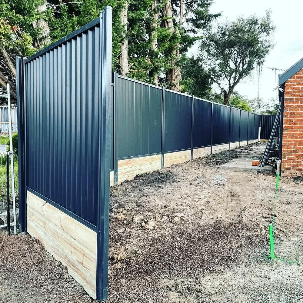A long, dark blue metal fence with vertical panels, installed on a concrete base, runs along a backyard with dirt ground and some green pipes lined up in the soil. There are trees and a brick building nearby.