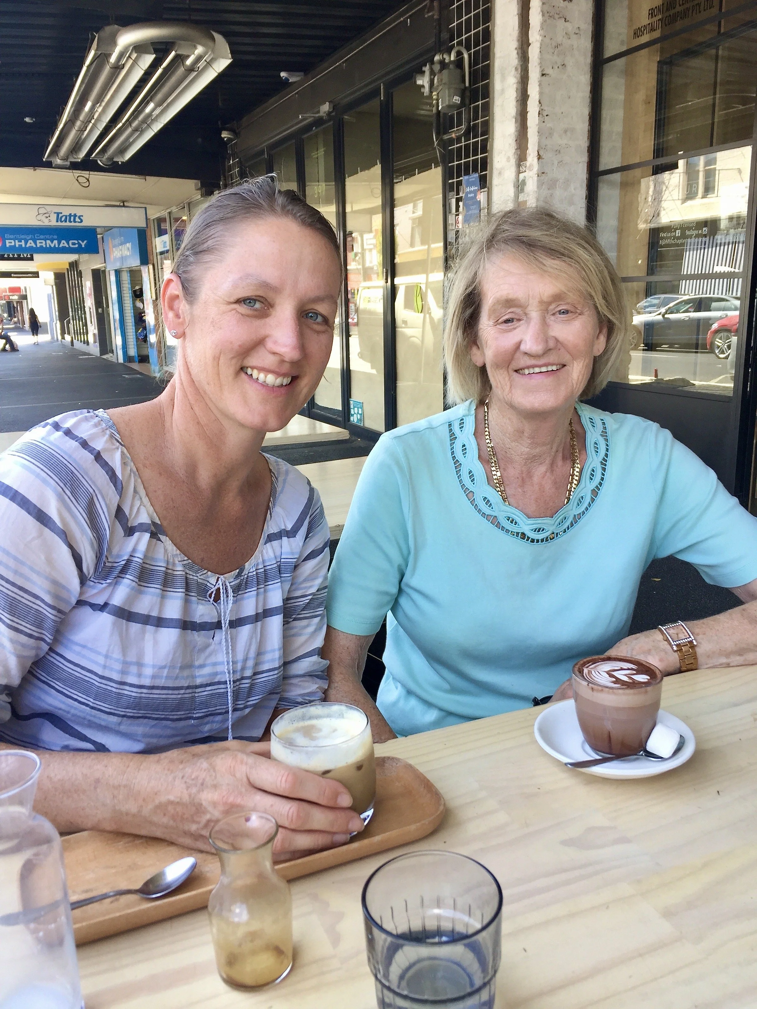 Two women sitting outdoors at a cafe table with various coffee drinks. One woman wears a striped shirt, and the other wears a light blue top with embroidery and a gold necklace.