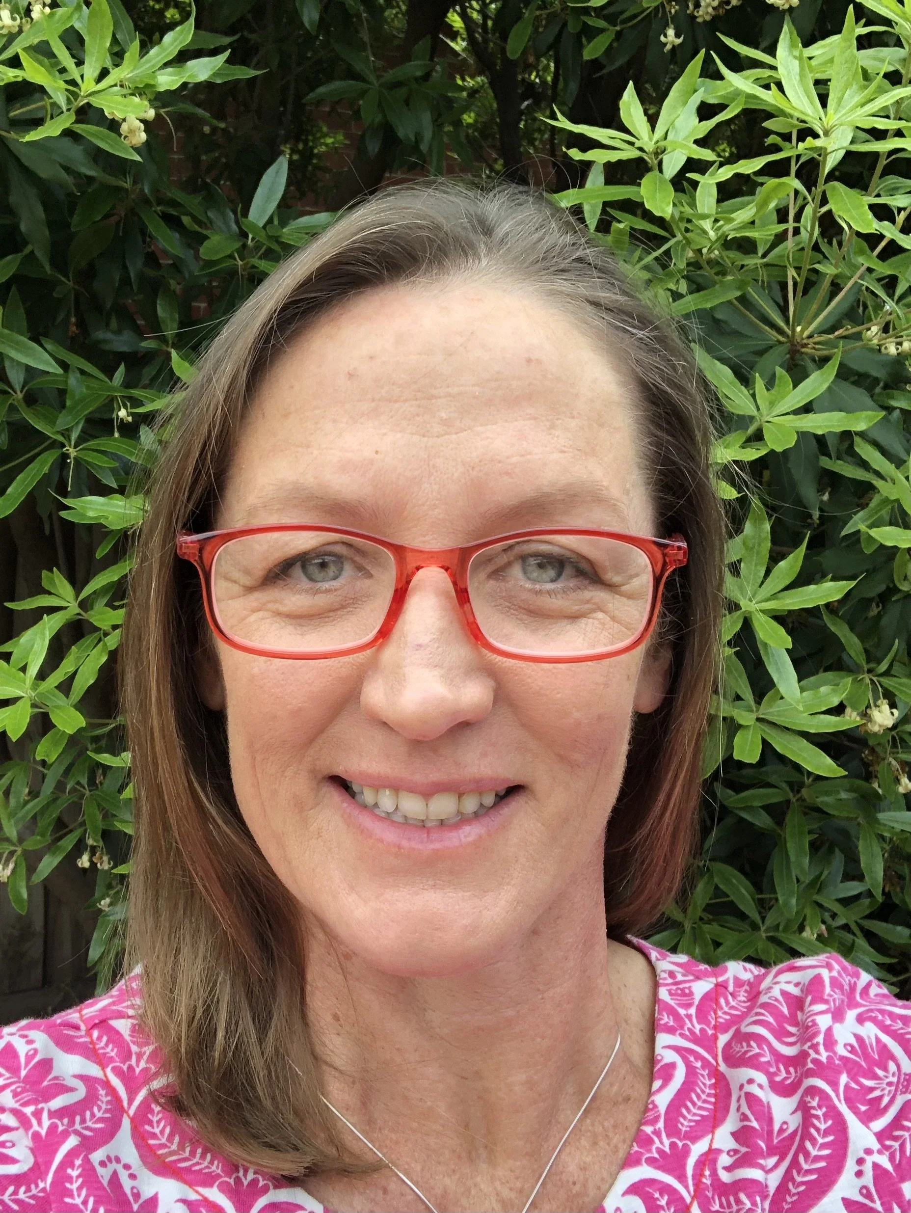 A woman with light skin, brown hair, and red glasses smiling outside. She is wearing a pink and white patterned top and a silver necklace. Green leafy bushes are in the background.