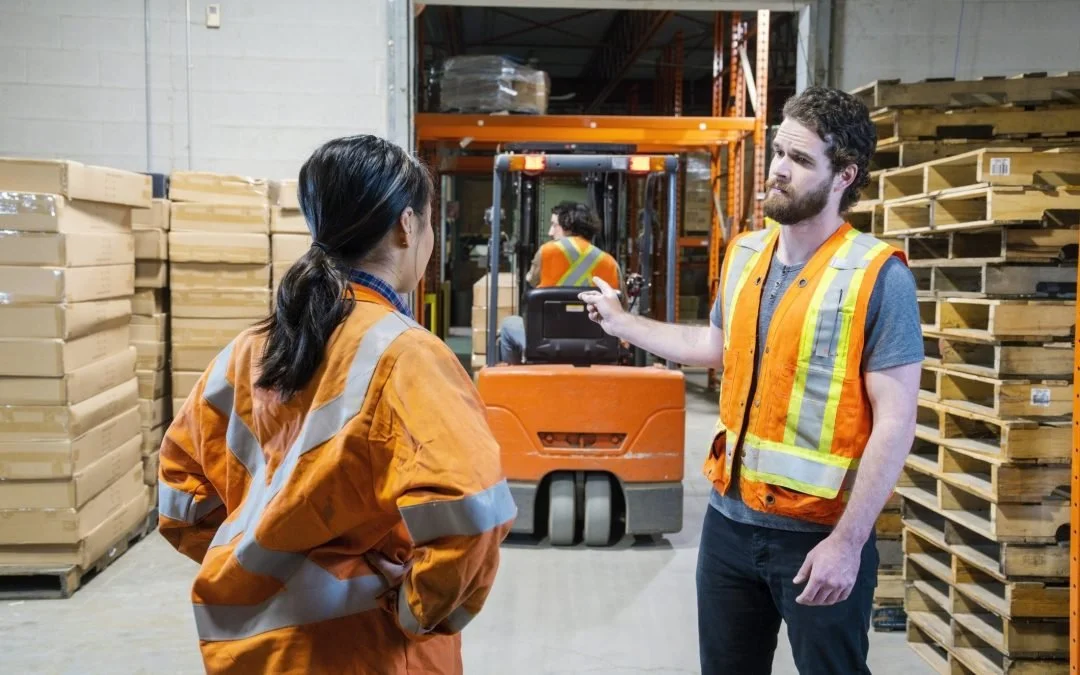 Two warehouse workers wearing high-visibility safety vests discussing workplace safety near a forklift in a storage facility.