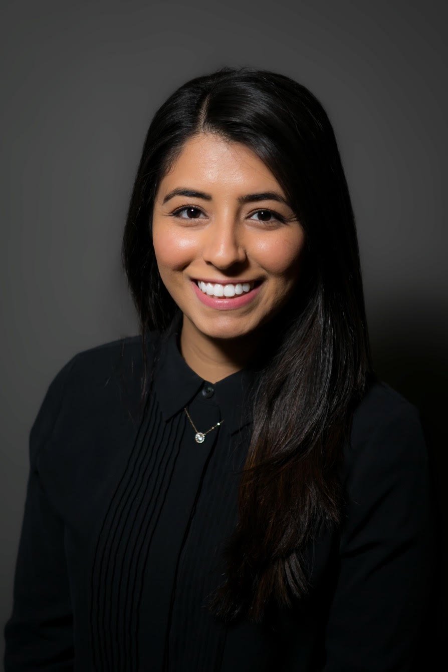 A young woman with long dark hair smiling, wearing a black button-up shirt and a delicate necklace, against a dark gray background.