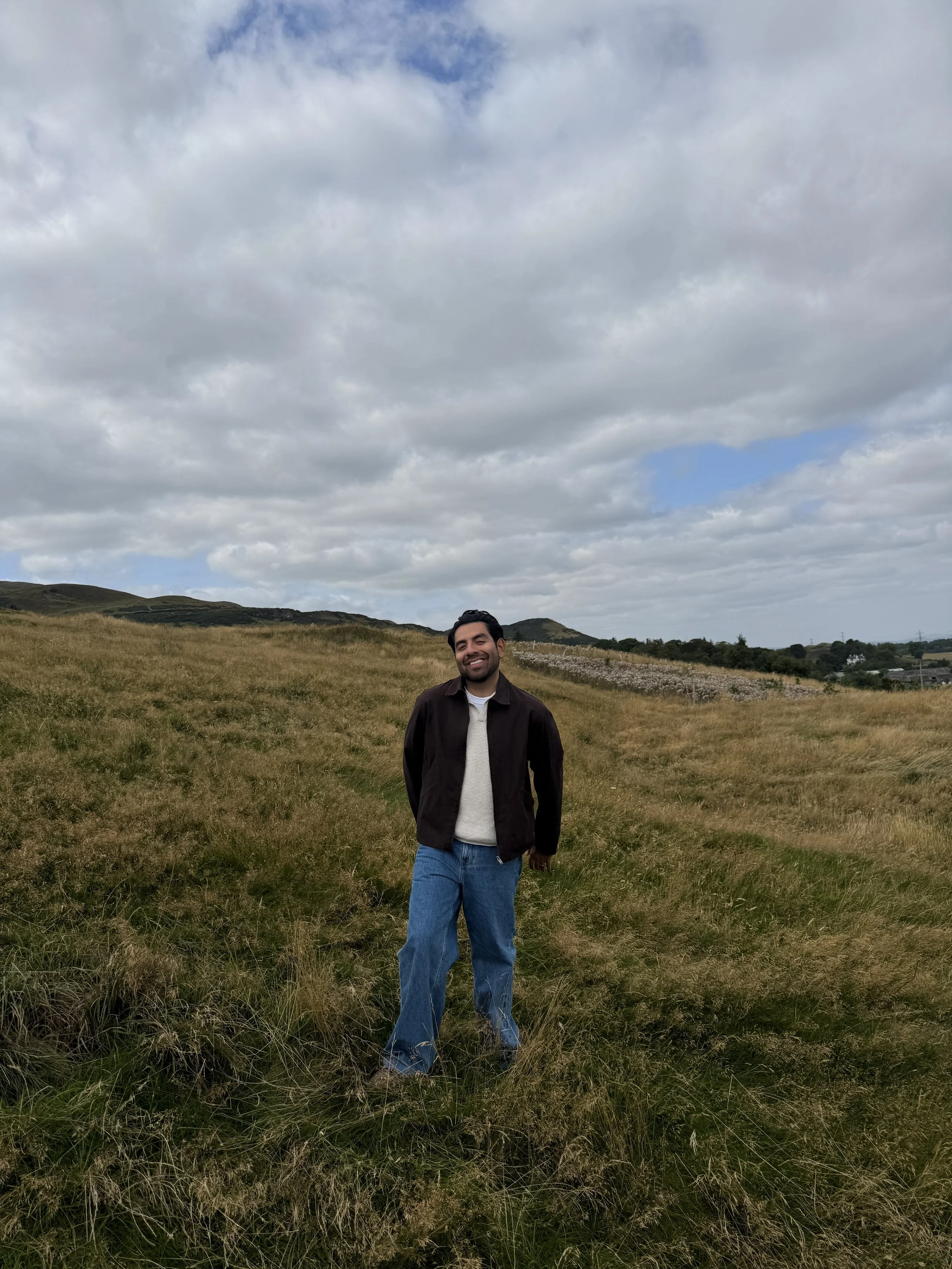 A man standing in a grassy field with hills and cloudy sky in the background.