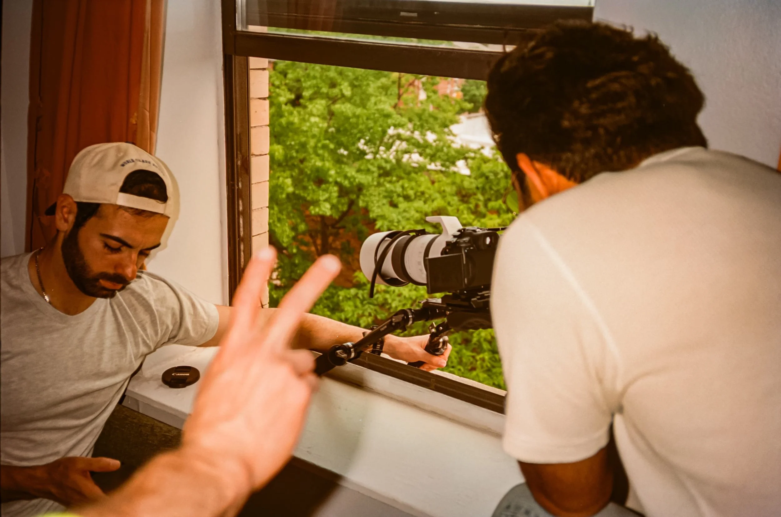 Two young men working with a camera on a window sill, one wearing a baseball cap and glasses, the other with curly hair, with trees visible outside the window.