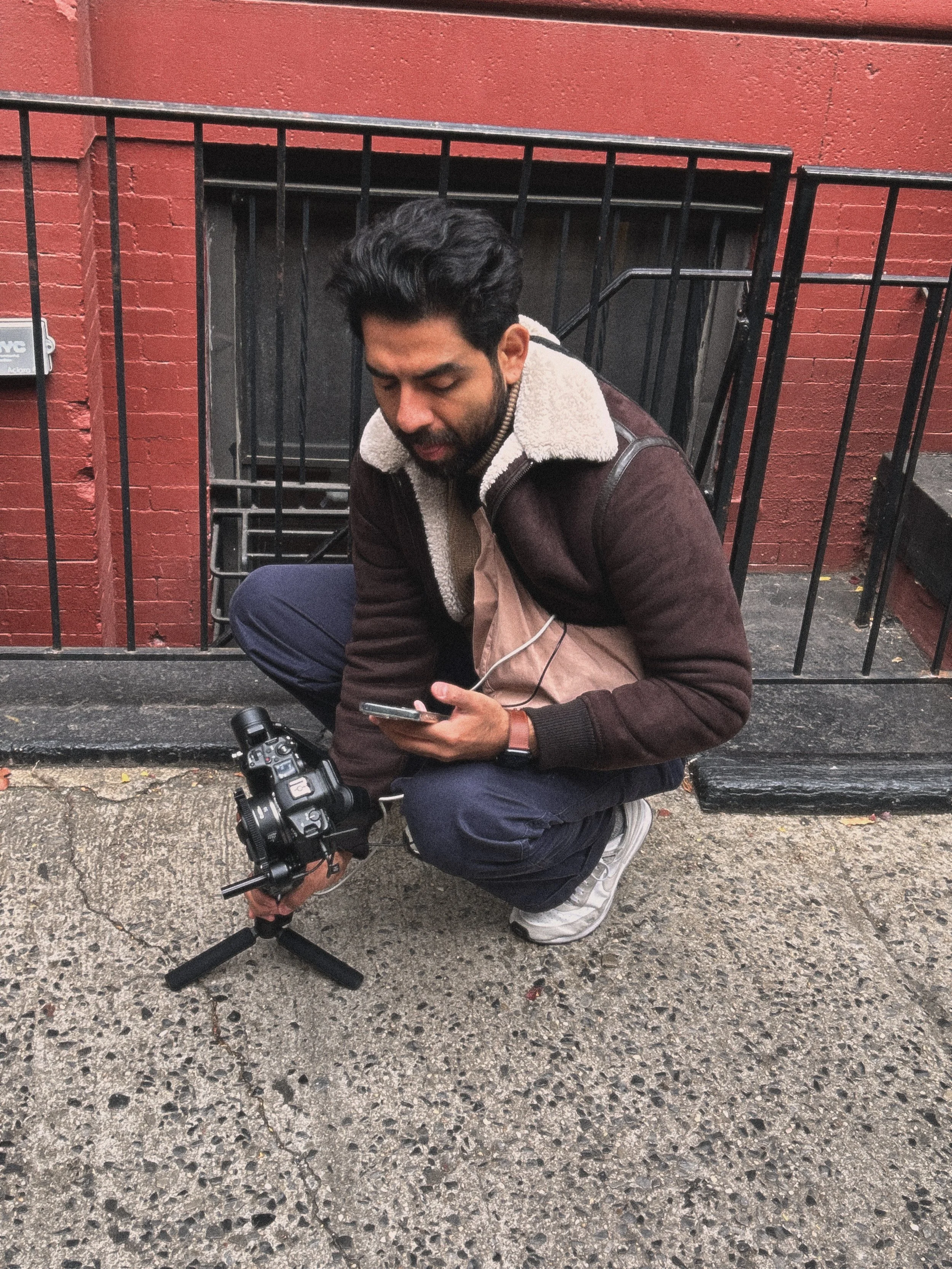 A man with dark hair and a beard is crouching on the sidewalk, holding a camera mounted on a tripod. He is looking at his phone, wearing a brown jacket with a shearling collar, blue pants, white sneakers, and a black watch. There is a red brick wall and black metal stairs in the background.