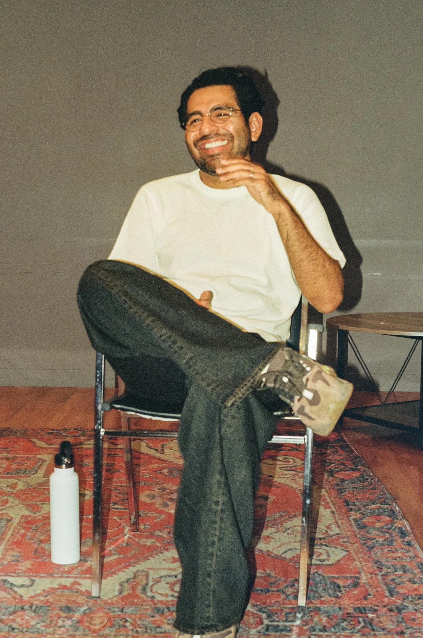 A man with glasses and a beard sitting on a chair with his legs crossed, smiling with his hand near his face, wearing a white t-shirt and camouflage sneakers, in a room with a patterned rug and a water bottle on the floor.