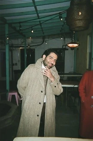 Man wearing a beige coat posing indoors with string lights and lamps in the background.