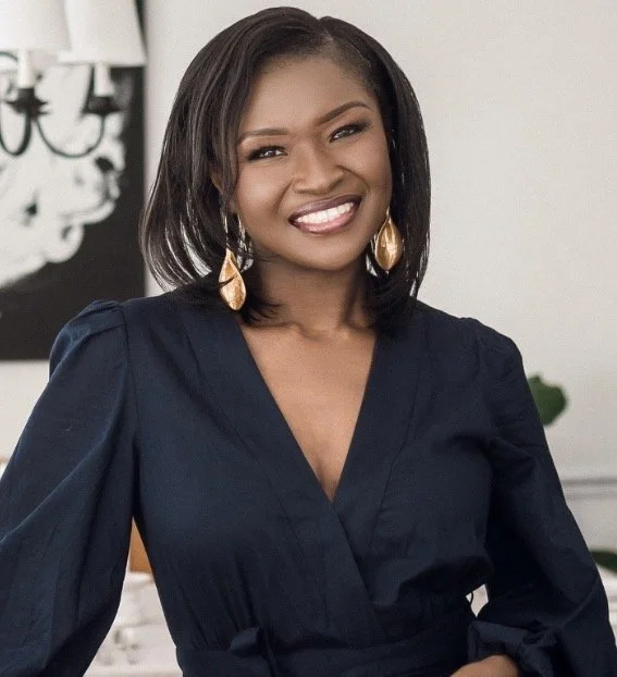 A smiling woman with shoulder-length black hair, wearing gold teardrop earrings and a navy blue dress, standing in a room with a white wall and a black and white painting in the background.