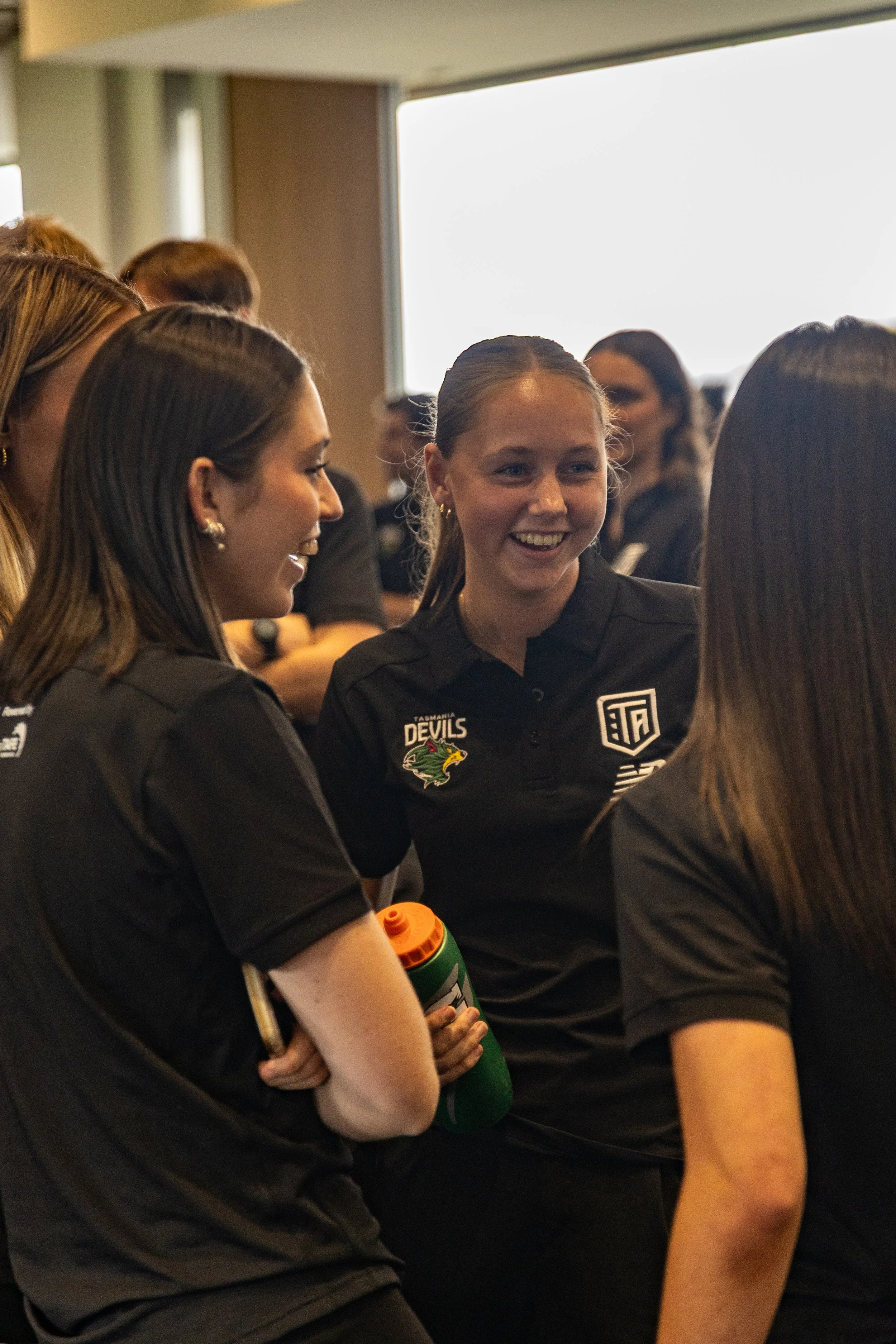 Smiling athletes in black Tasmania Devils team shirts gathered indoors, with one holding a sports water bottle.