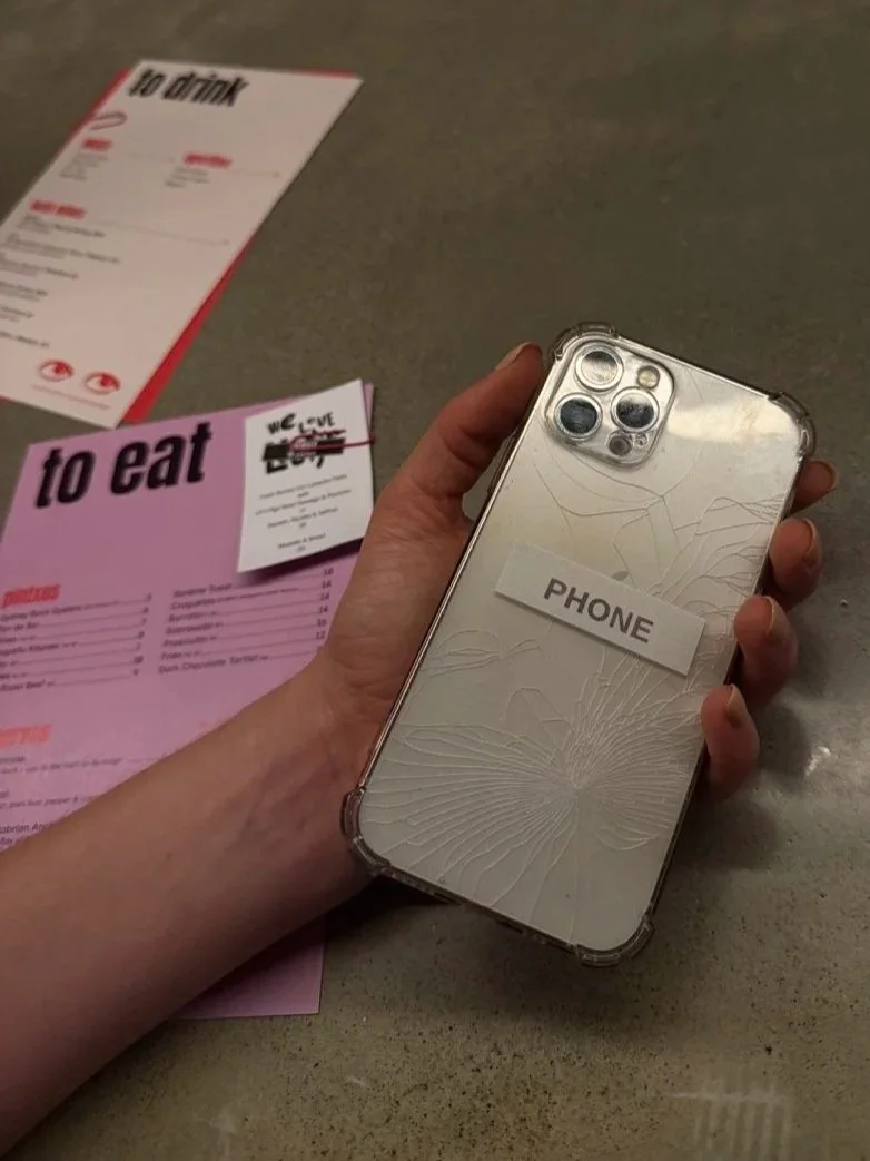A person holding a cracked phone with a label that says 'PHONE' on it, on a table with menus in the background.