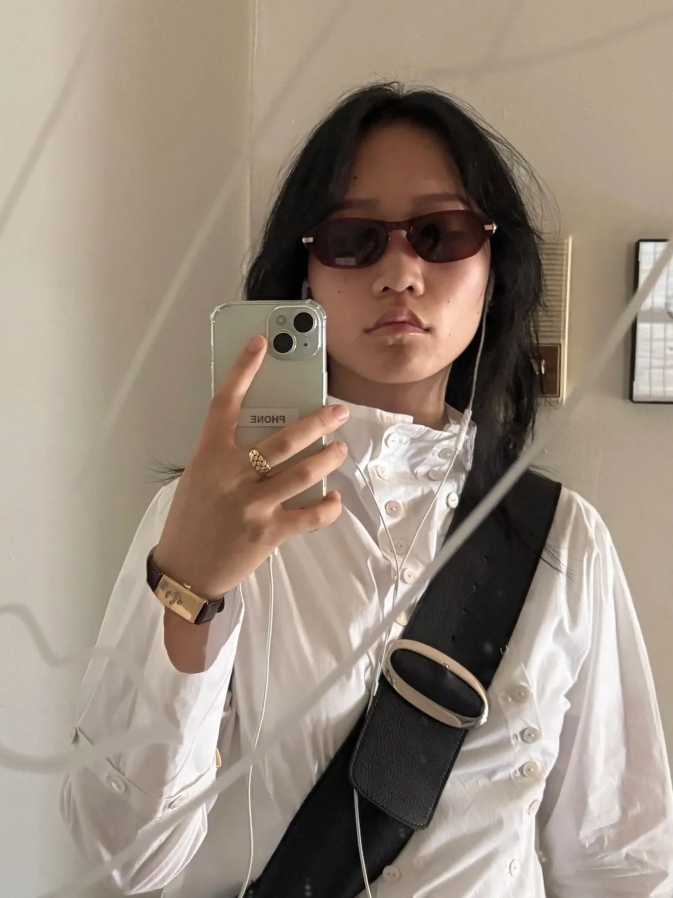 A woman taking a selfie in a mirror, wearing sunglasses, a white button-up shirt, and a black strap across her chest, with a gold ring and a gold watch, holding an iPhone with the label that says 'PHONE' on it.