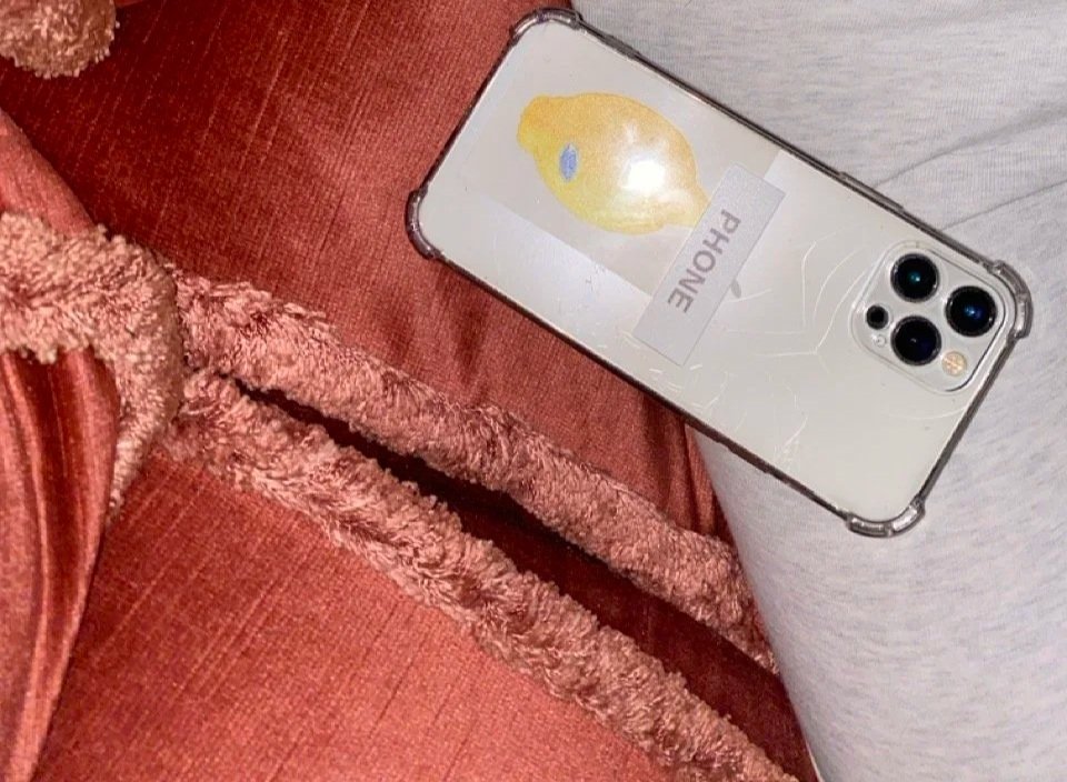 Close-up of a smartphone with a cracked screen, and with the label that says 'PHONE' on it resting on a person's lap, who is wearing gray clothing and sitting on a reddish textured fabric surface.