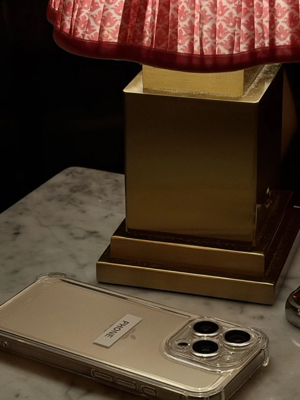 A gold and pink lamp on a marble surface with a white iPhone with three camera lenses and a label reading 'PHONE' in black letters in front.