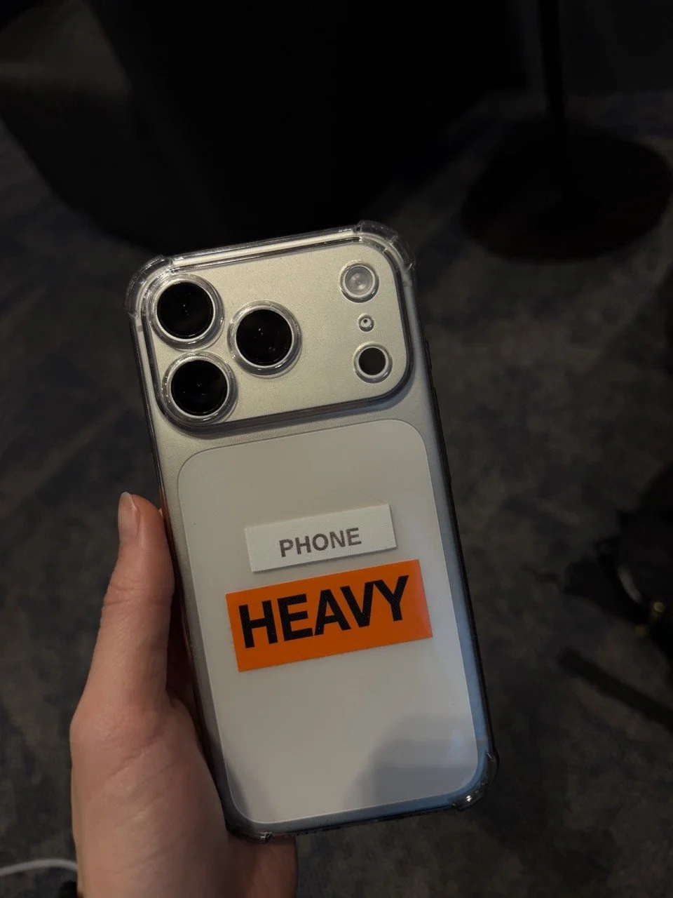 Hand holding a silver iPhone with a transparent case, labeled 'PHONE HEAVY', against a dark background.