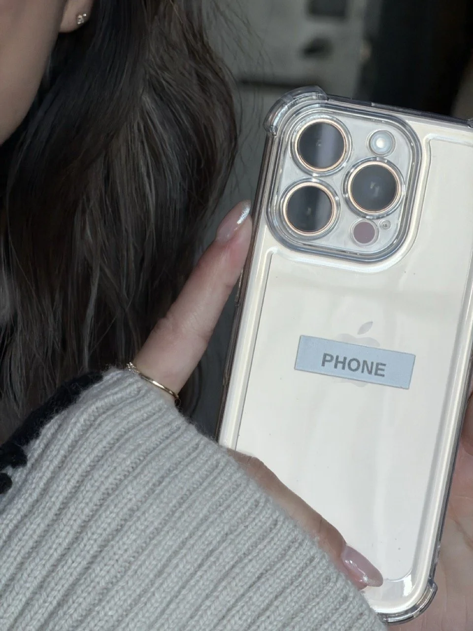 A person holding a white iPhone with a clear case that has a sticker labeled 'PHONE' on the back, showing the camera lenses on the top left corner.