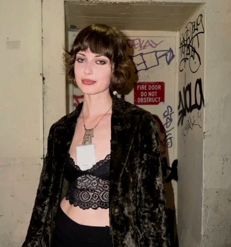 A young woman with short brown hair and makeup, wearing a black lace crop top, and a necklace with a large pendant, standing in front of a graffiti-covered wall and a red fire door. A phone near her chest has a label that says 'PHONE' on it.