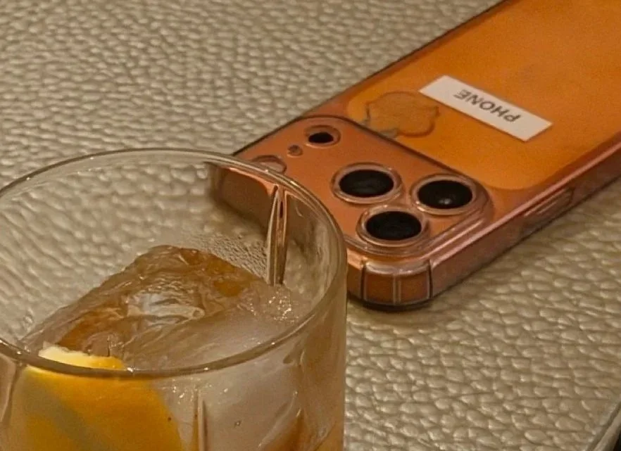 A clear glass with ice cubes and a lemon wedge on a textured surface, with a rose gold phone in the background labeled with 'PHONE'.