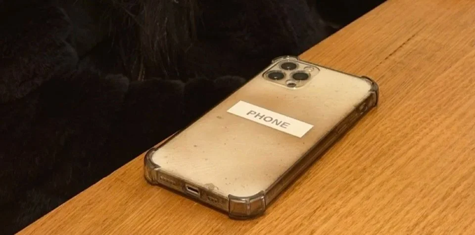 A dusty, scratched iPhone with a label that says 'PHONE' rests on a wooden table.