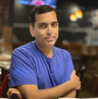 A man in a blue shirt sitting at a wooden table in a restaurant or cafe.