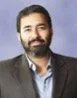 A man with a beard and dark hair dressed in a dark blazer and light blue shirt, against a blue gradient background.