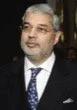 A man with gray hair and glasses wearing a dark suit and tie, standing indoors.