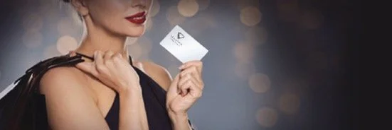A woman in a black dress holding a small white card with a logo, with a blurred bokeh background.