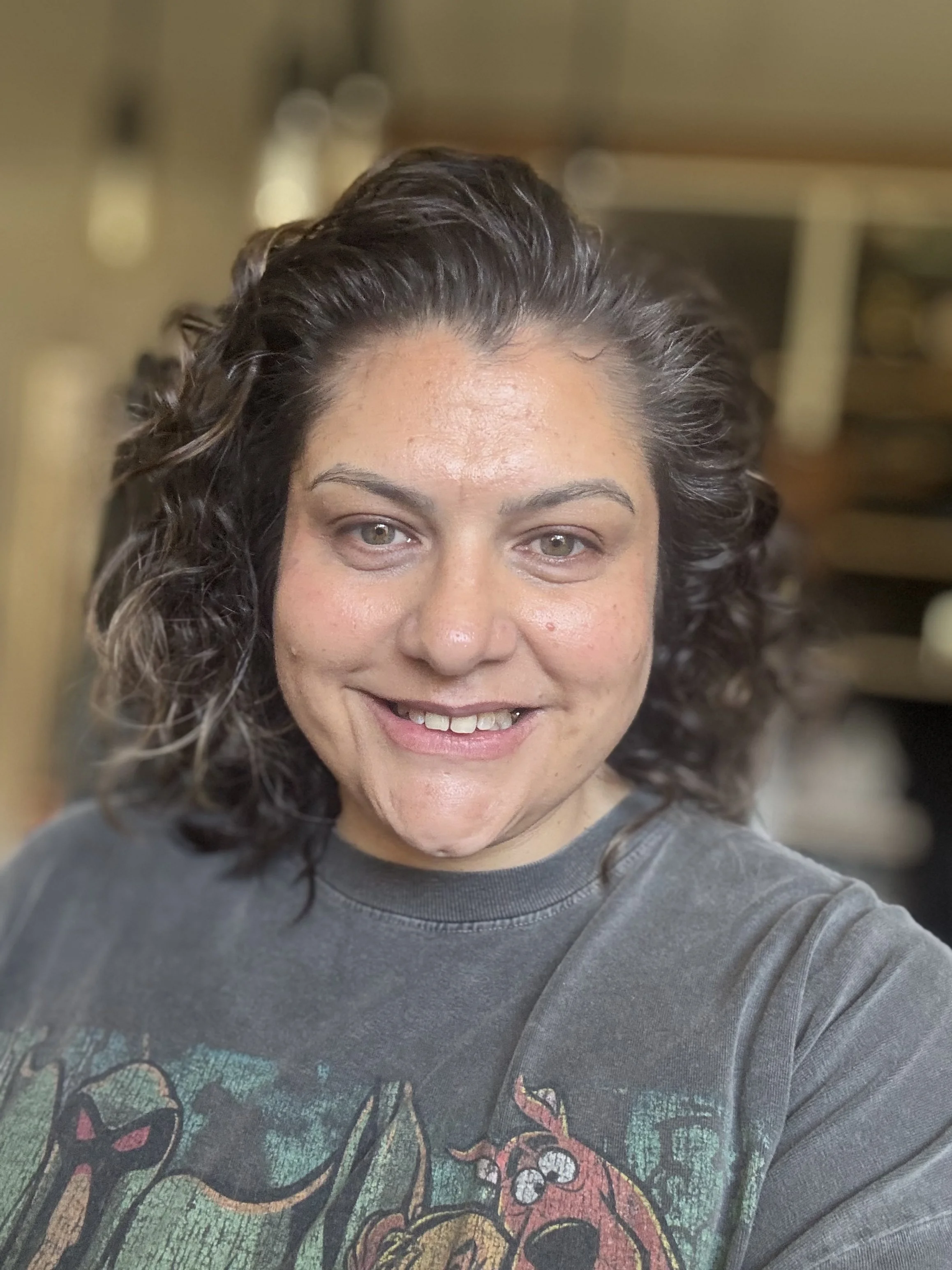Smiling woman with curly dark hair wearing a gray graphic T-shirt, photographed indoors.