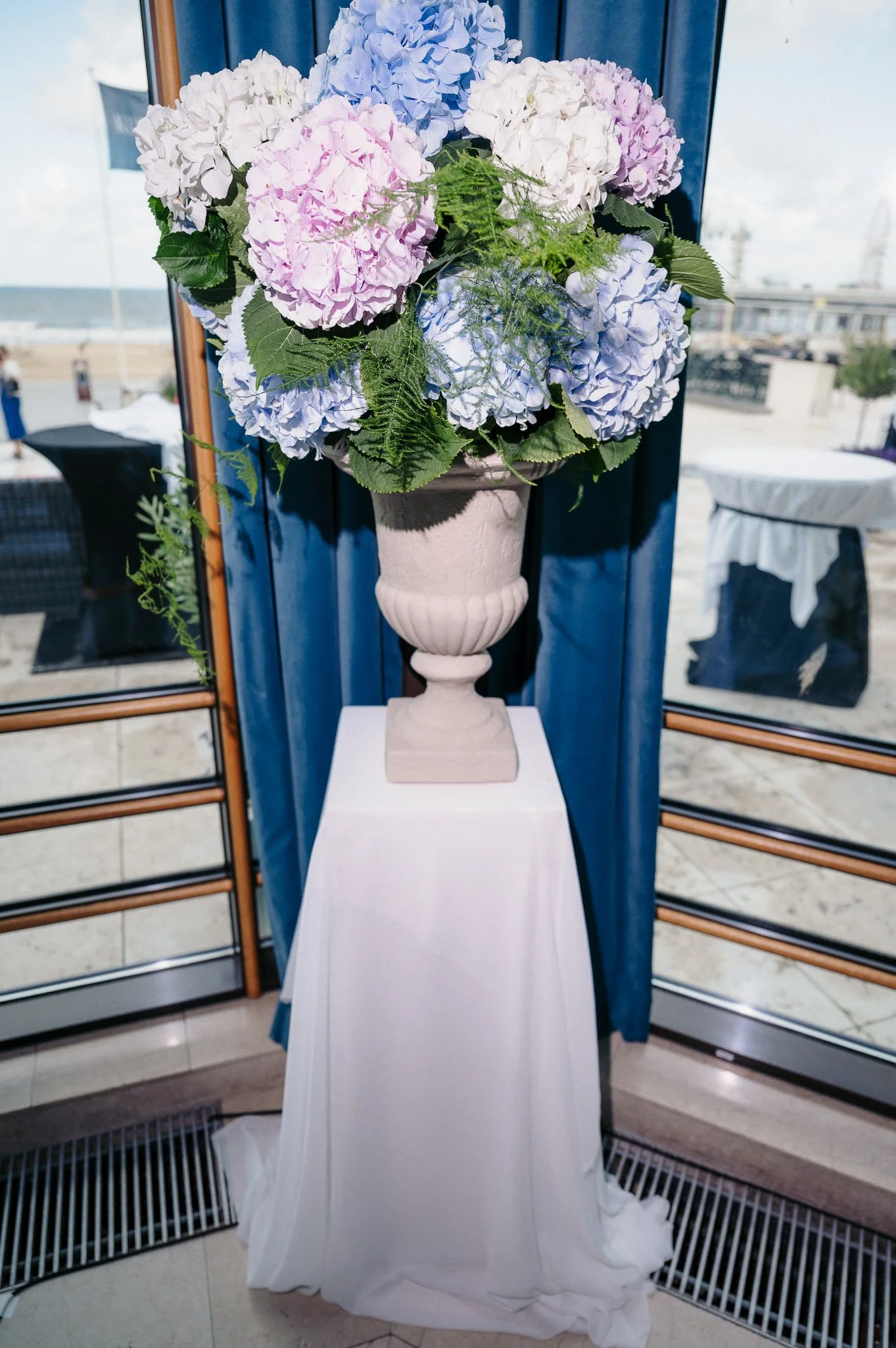 Pastel blue, pink, and white hydrangea arrangement in an ornate vase on a white-draped pedestal by a seaside window.