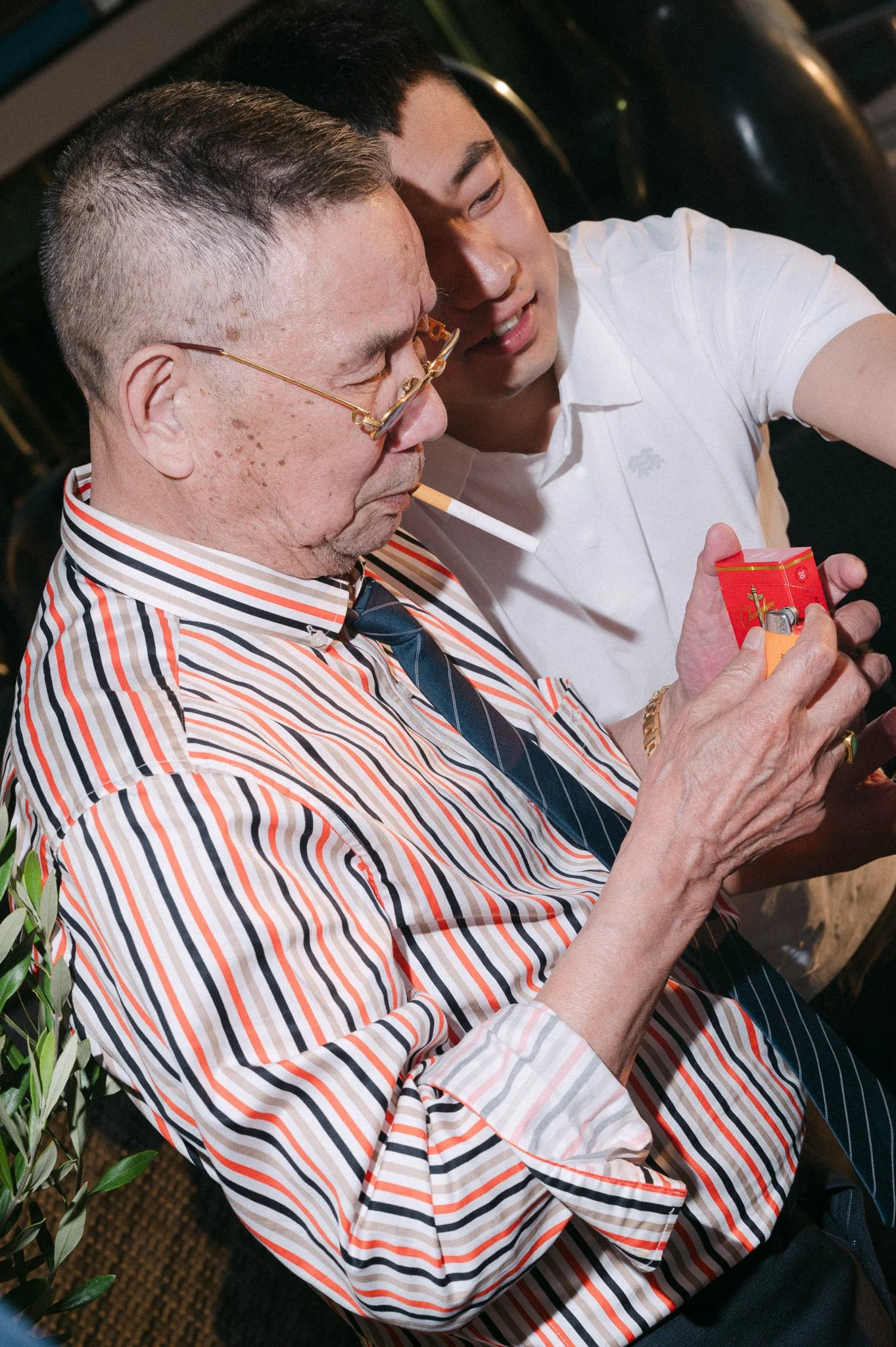 Two men sit closely together; one wears glasses and a striped shirt while holding a cigarette and red cigarette pack, and the other wears a white polo shirt.