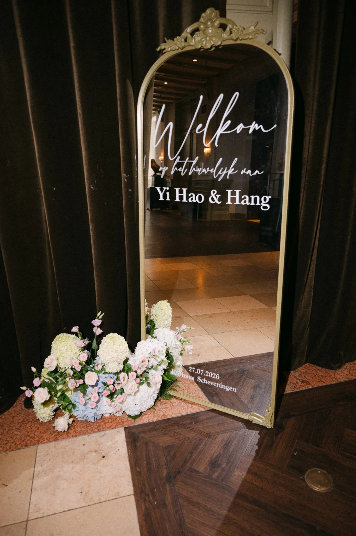 Ornate gold mirror wedding sign reading “Welkom,” “Yi Hao & Hang,” and “27.07.2026 Kurhaus Scheveningen,” decorated with pastel flowers.