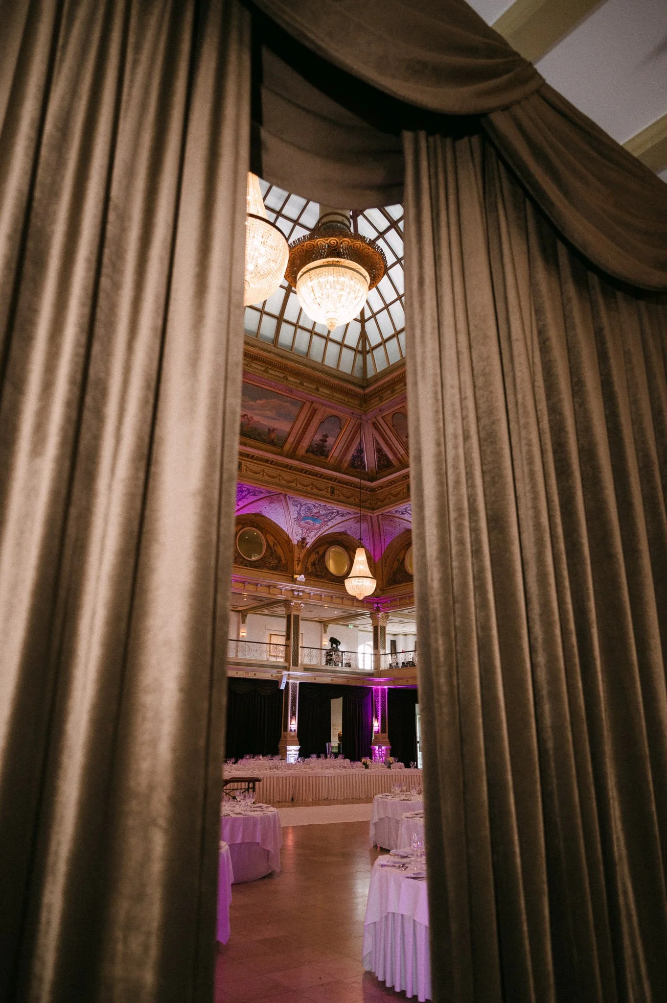 Elegant banquet hall viewed through velvet curtains, featuring chandeliers, a glass-domed ceiling, ornate murals, and round dining tables.