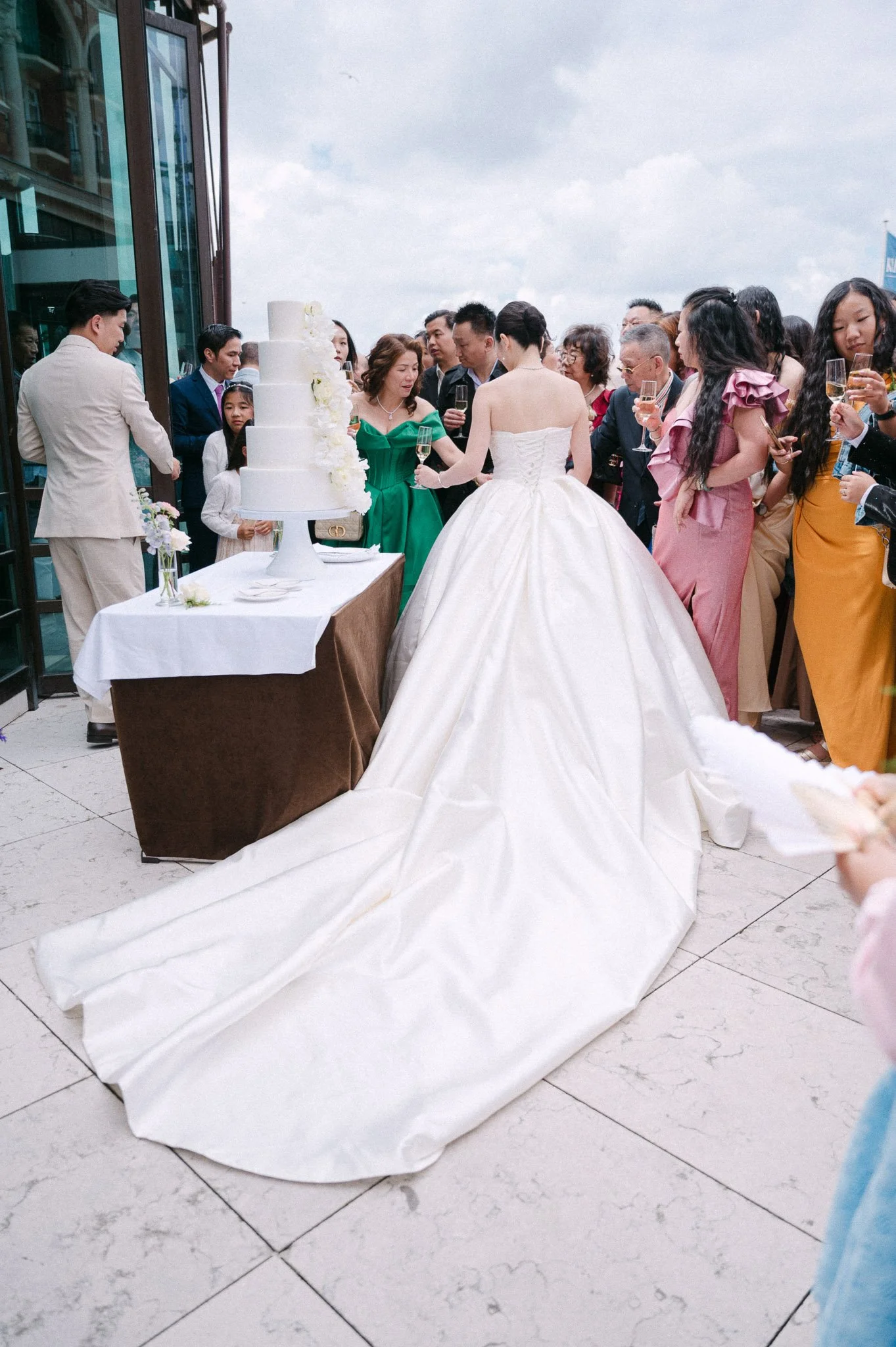 Bride in a long white wedding gown surrounded by guests holding champagne near a multi-tiered wedding cake.