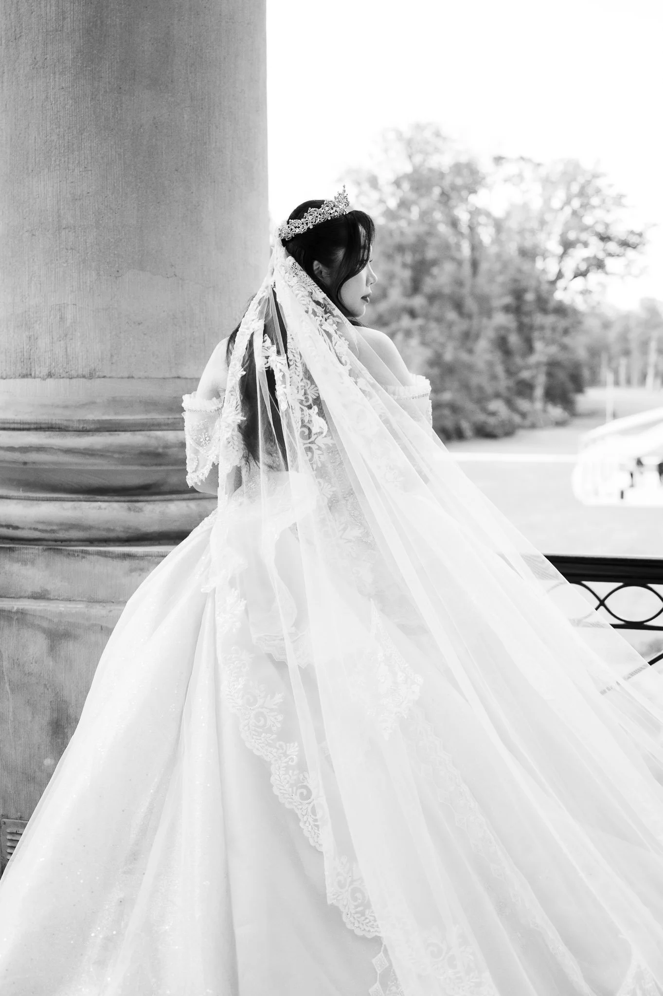Black-and-white portrait of a bride in a tiara and lace veil beside a stone column.