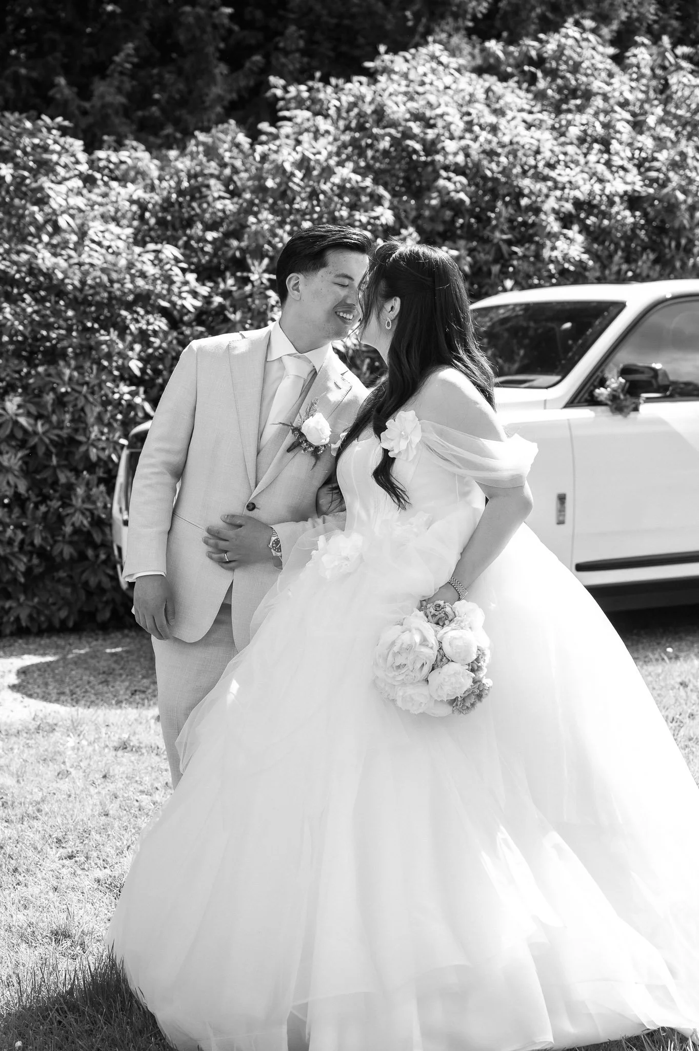 Newlywed couple embracing outdoors beside a white wedding car, with the bride holding a bouquet.