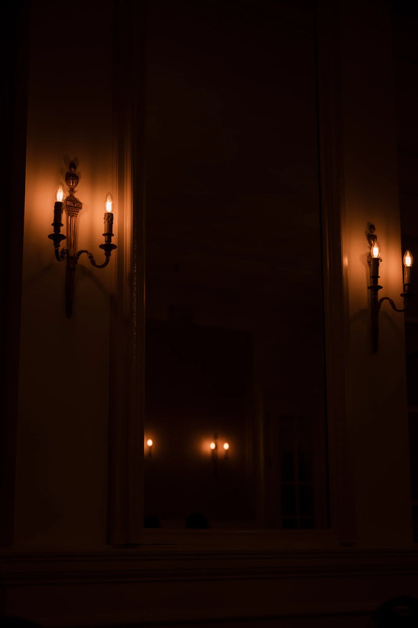 Dimly lit ornate wall sconces glowing beside a dark window, creating a warm vintage interior atmosphere