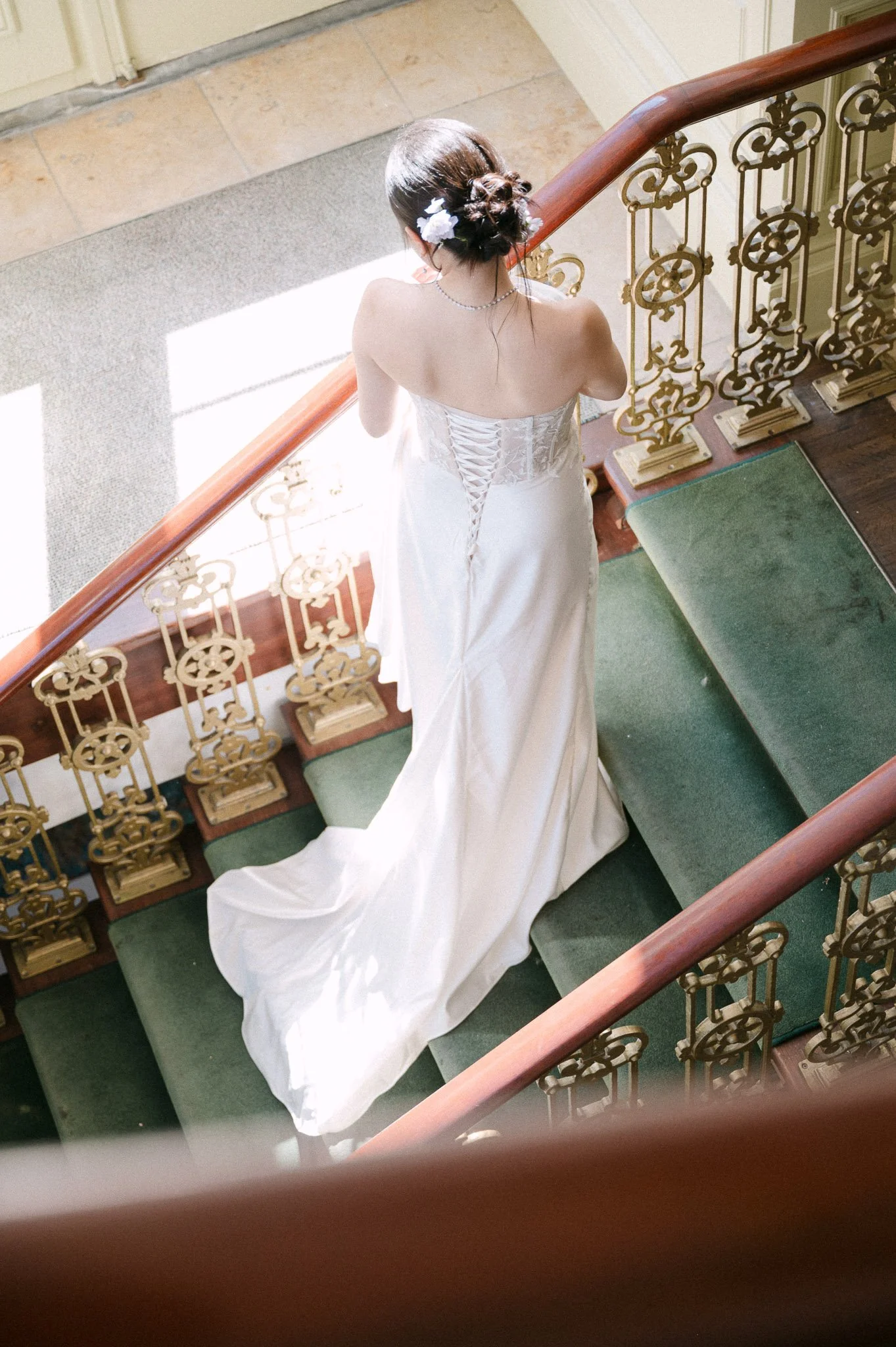 Bride in a white wedding gown with floral hair accessories walking down a green-carpeted staircase with ornate railings