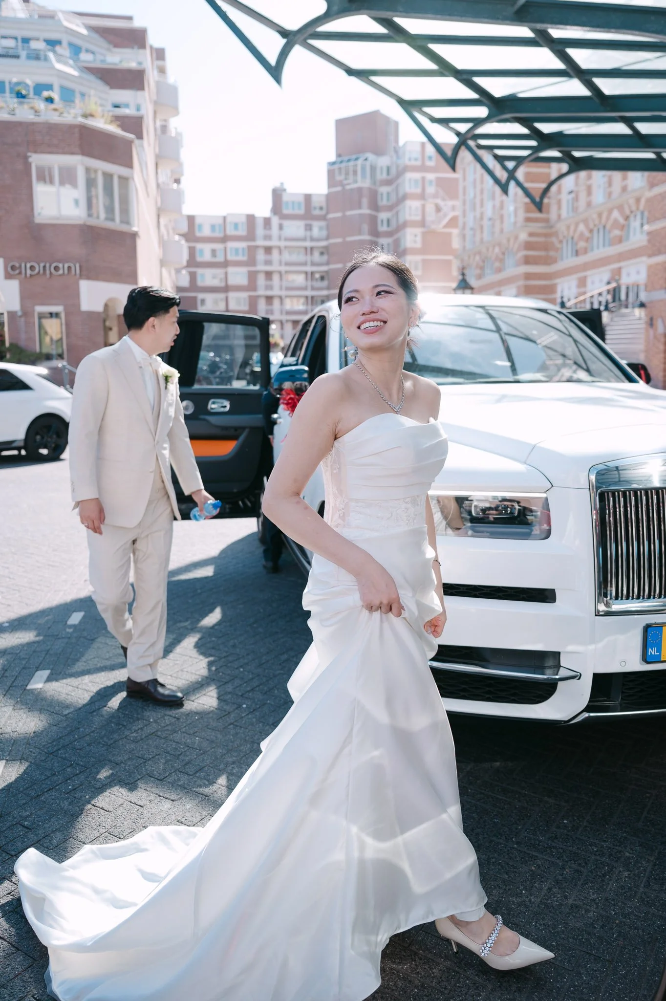 Smiling bride in a white wedding gown standing beside a white luxury car, with the groom behind her.