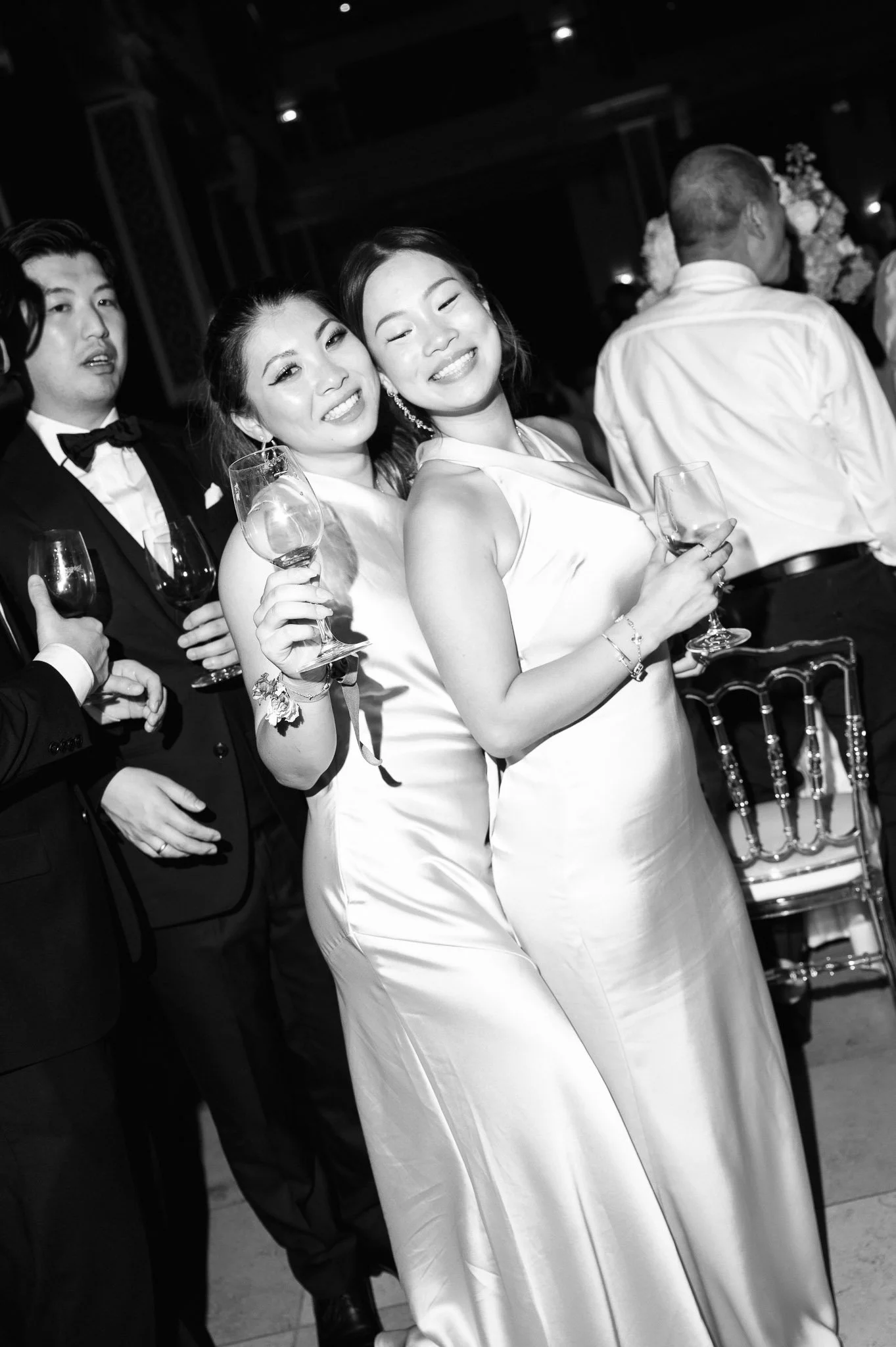 Black-and-white wedding reception photo of two smiling women in white dresses holding wine glasses, with guests in formalwear behind them.