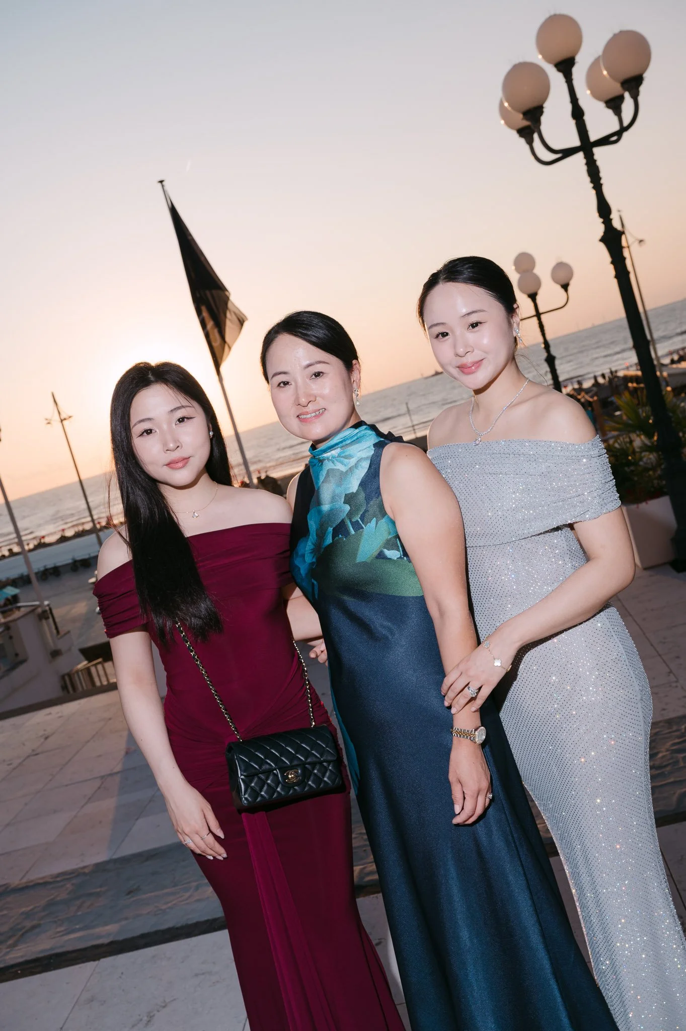 Three women in elegant evening dresses pose by the seaside at sunset, with a flag and globe-topped street lamps behind them.