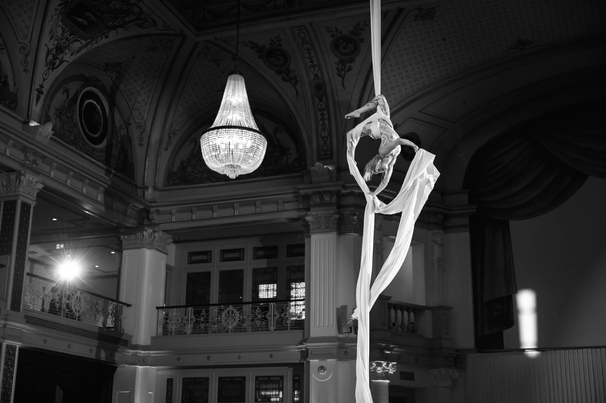 Aerial silk dancer performing in an ornate theater beneath a crystal chandelier.