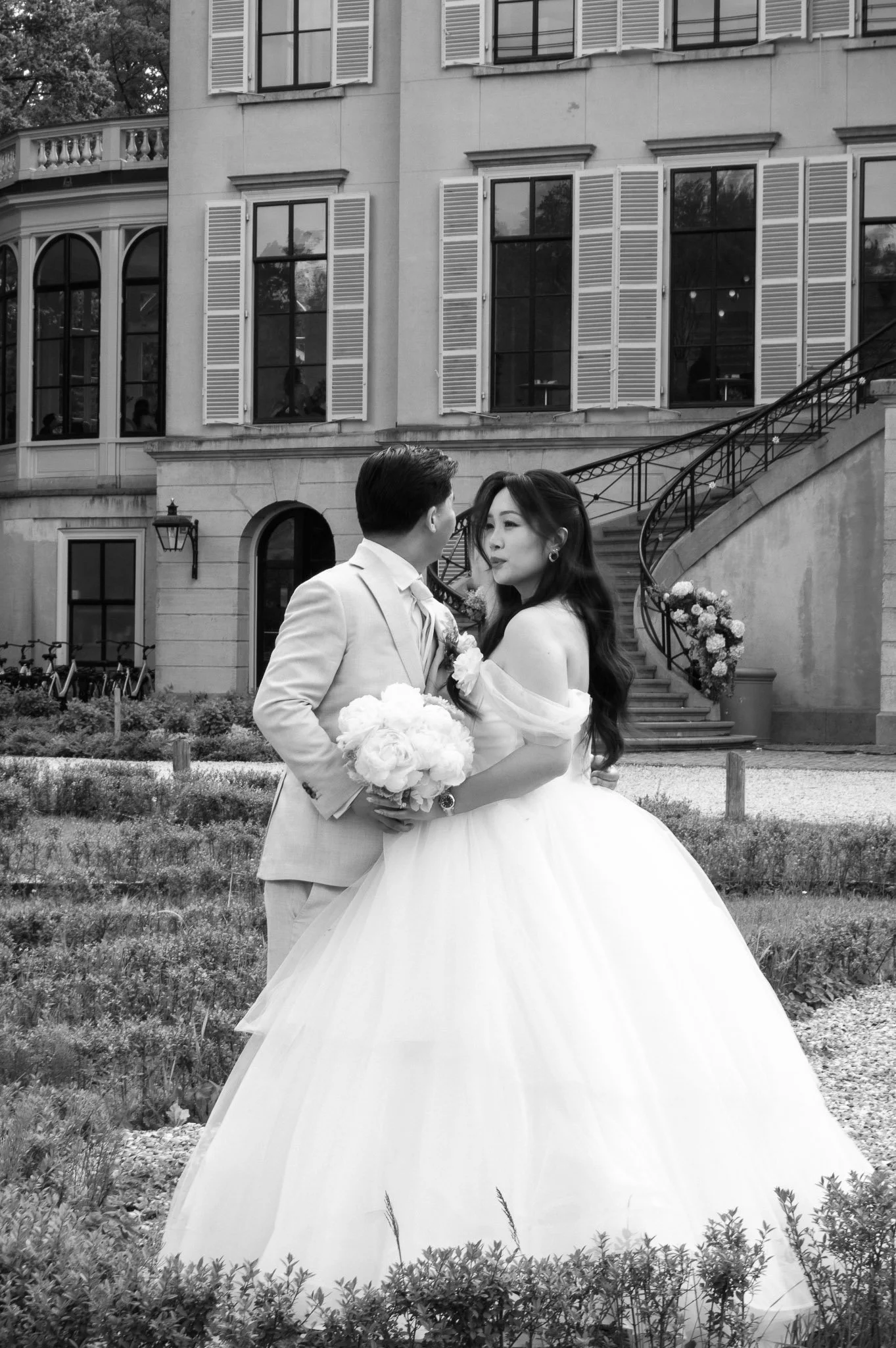 Black-and-white wedding portrait of a bride holding a bouquet with her groom in front of a stately mansion and garden staircase