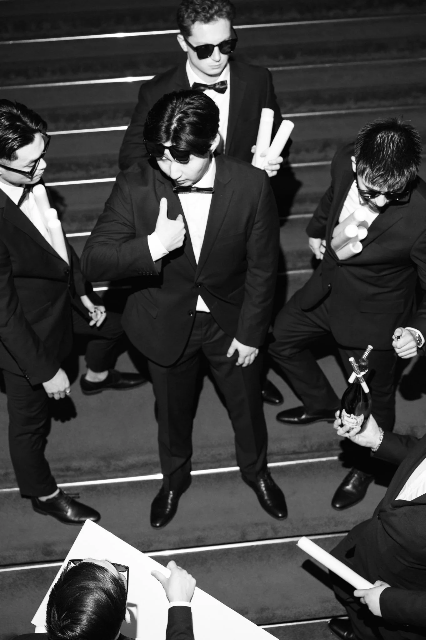 Black-and-white photo of four men in tuxedos and sunglasses holding rolled papers at a formal event.