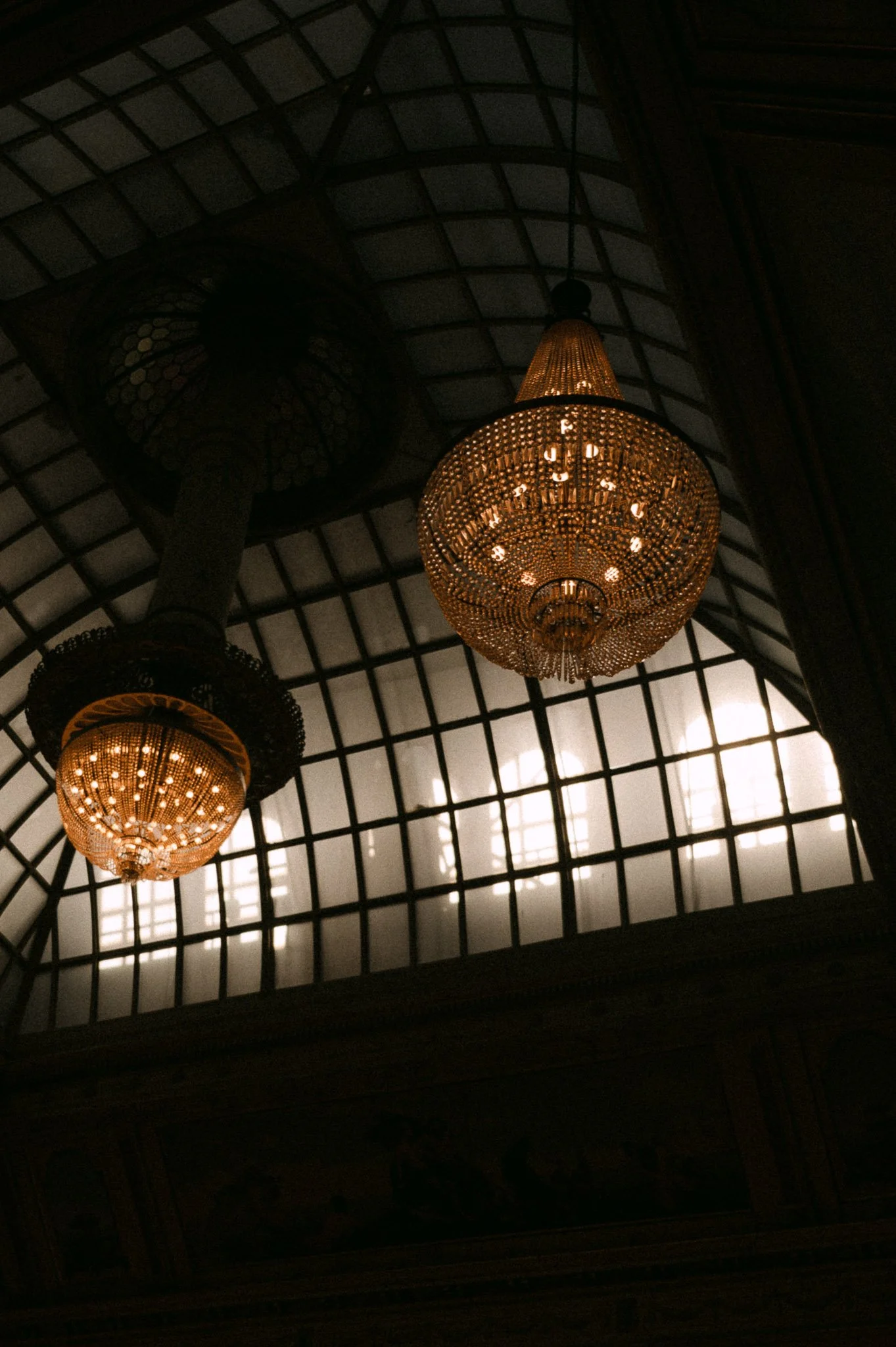 Two ornate illuminated chandeliers hanging beneath a vaulted glass-and-metal ceiling