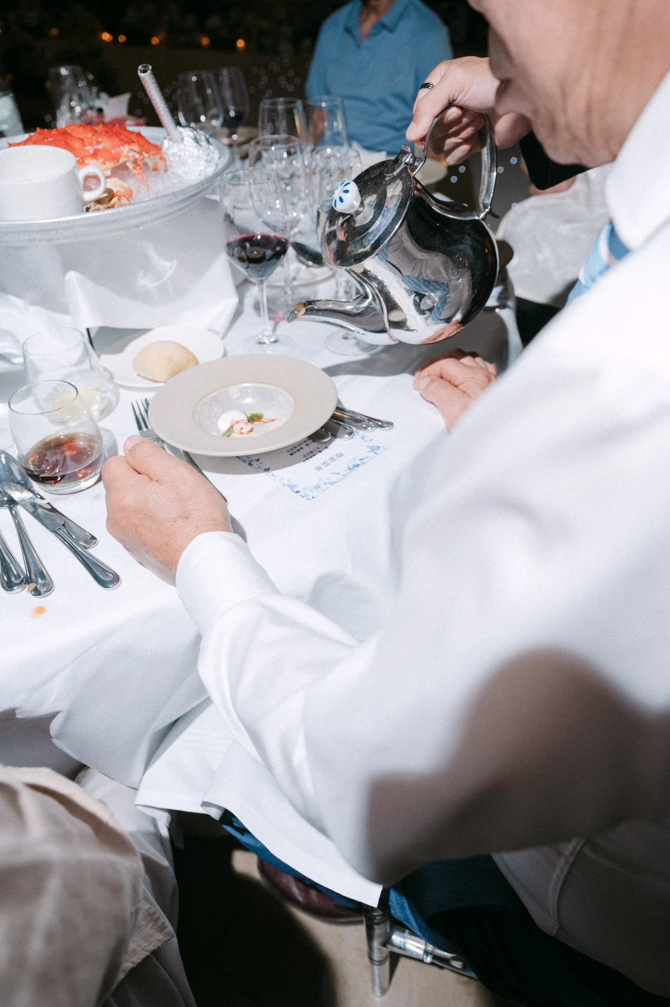 Server pouring water from a silver pitcher at an elegant restaurant table with wine glasses, fine china, and lobster.