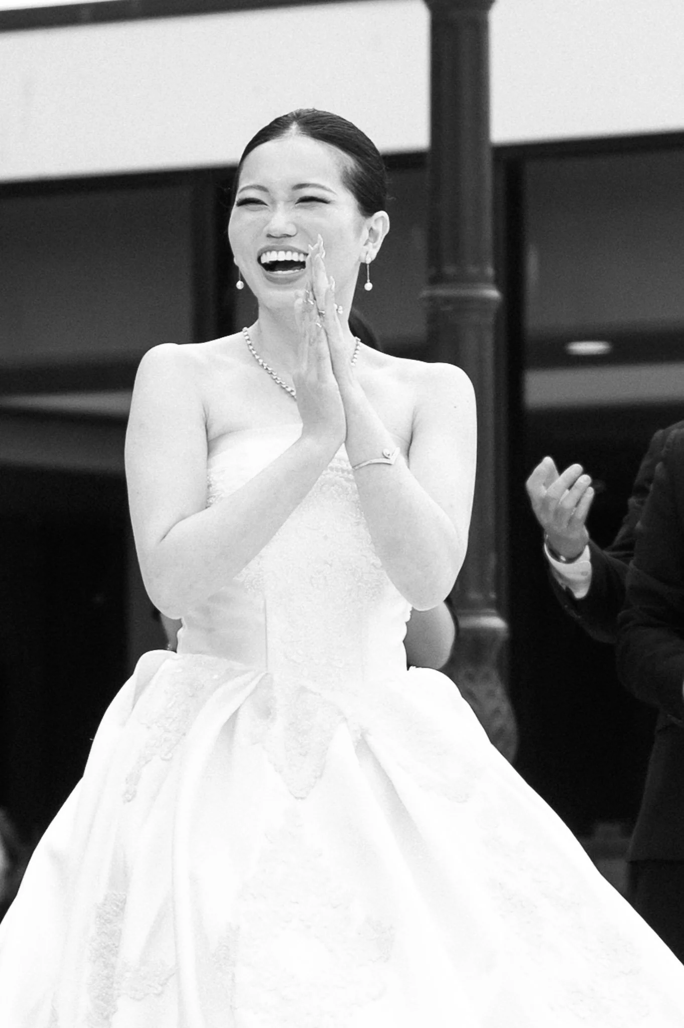 Black-and-white photo of a smiling woman in a strapless wedding gown clapping at an event.