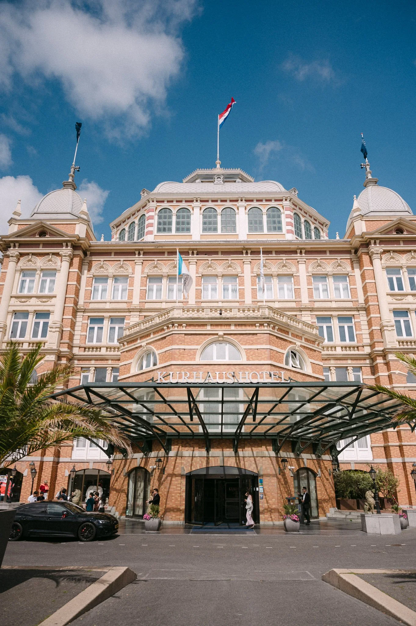 Grand Kurhaus Hotel entrance with ornate brick architecture, flags, glass canopy, and palm trees under a blue sky